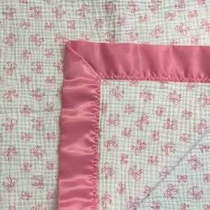 May include: A white baby blanket with a pink bow pattern and a pink satin border. The blanket is made of a soft, textured fabric, perfect for a nursery or baby shower gift.