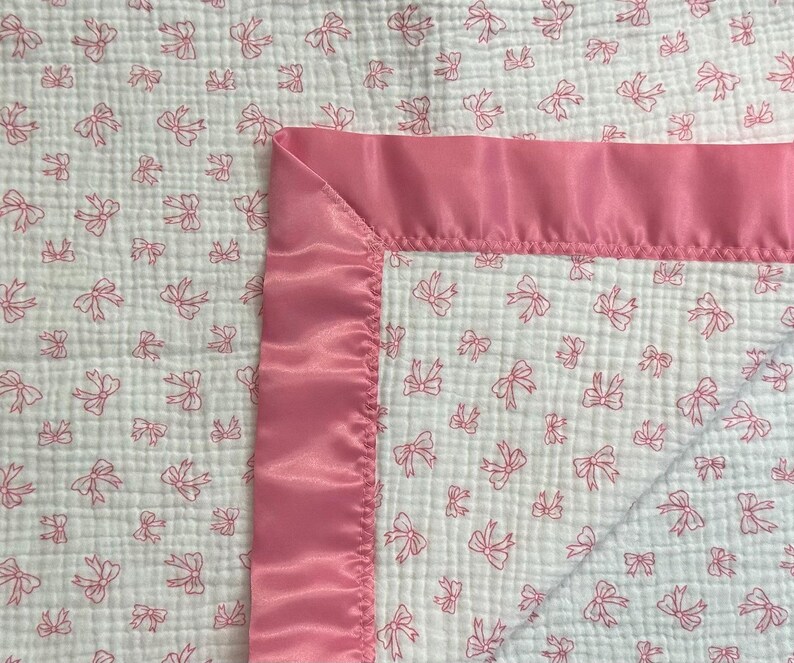 May include: A white baby blanket with a pink bow pattern and a pink satin border. The blanket is made of a soft, textured fabric, perfect for a nursery or baby shower gift.
