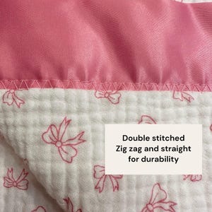 May include: Close-up of a pink and white baby blanket. The top edge is a solid pink satin-like fabric. The lower portion is white with a pink bow pattern. The text on the image reads: "Double stitched, Zig zag and straight for durability."