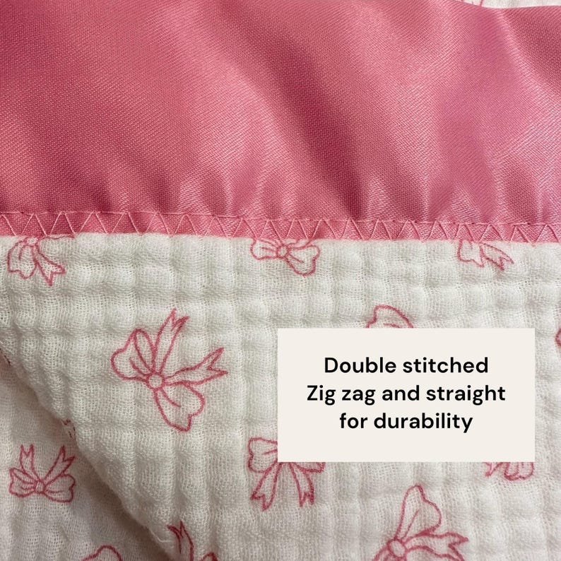 May include: Close-up of a pink and white baby blanket. The top edge is a solid pink satin-like fabric. The lower portion is white with a pink bow pattern. The text on the image reads: "Double stitched, Zig zag and straight for durability."