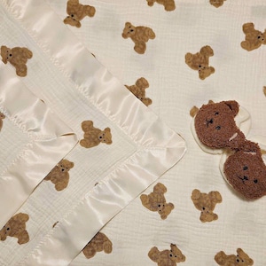May include: A white muslin baby blanket with a satin trim and a brown teddy bear print. The blanket is folded in thirds and there are two brown teddy bear plush toys on top of the blanket.