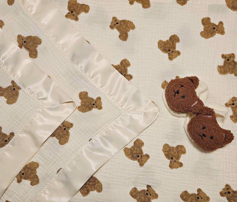 May include: A white muslin baby blanket with a satin trim and a brown teddy bear print. The blanket is folded in thirds and there are two brown teddy bear plush toys on top of the blanket.