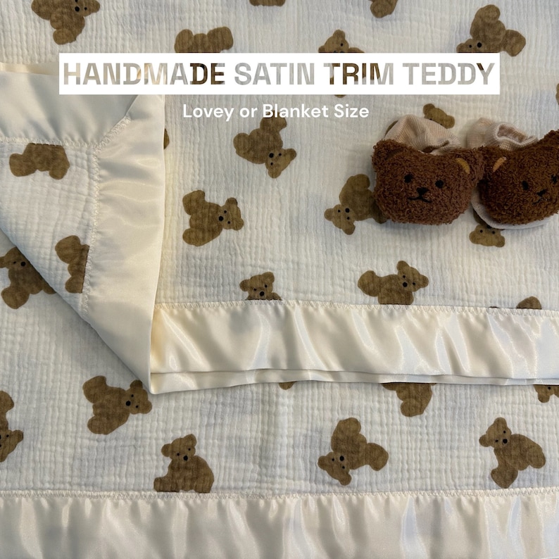 May include: A white muslin baby blanket with a satin trim and a brown teddy bear print. The blanket is folded in half and has a satin trim around the edges. The blanket is labeled "HANDMADE SATIN TRIM TEDDY Lovey or Blanket Size".