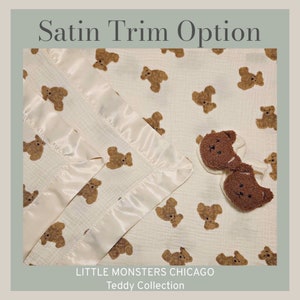 May include: A white muslin baby blanket with a satin trim and a brown teddy bear print. The blanket is folded in half and has a pair of brown teddy bear booties on top. The text "LITTLE MONSTERS CHICAGO Teddy Collection" is printed on the bottom of the blanket.