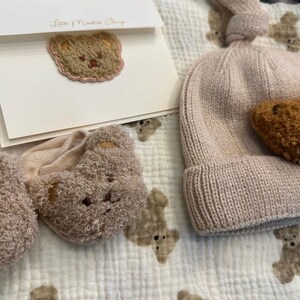 May include: A set of baby clothes with a brown bear theme. The set includes a knitted hat with a brown bear applique, a pair of socks with brown bear faces, and a card with a brown bear embroidered on it. The card says "Love & Nurture Always".