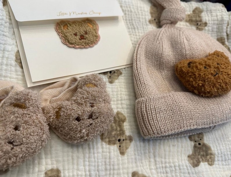 May include: A set of baby clothes with a brown bear theme. The set includes a knitted hat with a brown bear applique, a pair of socks with brown bear faces, and a card with a brown bear embroidered on it. The card says "Love & Nurture Always".