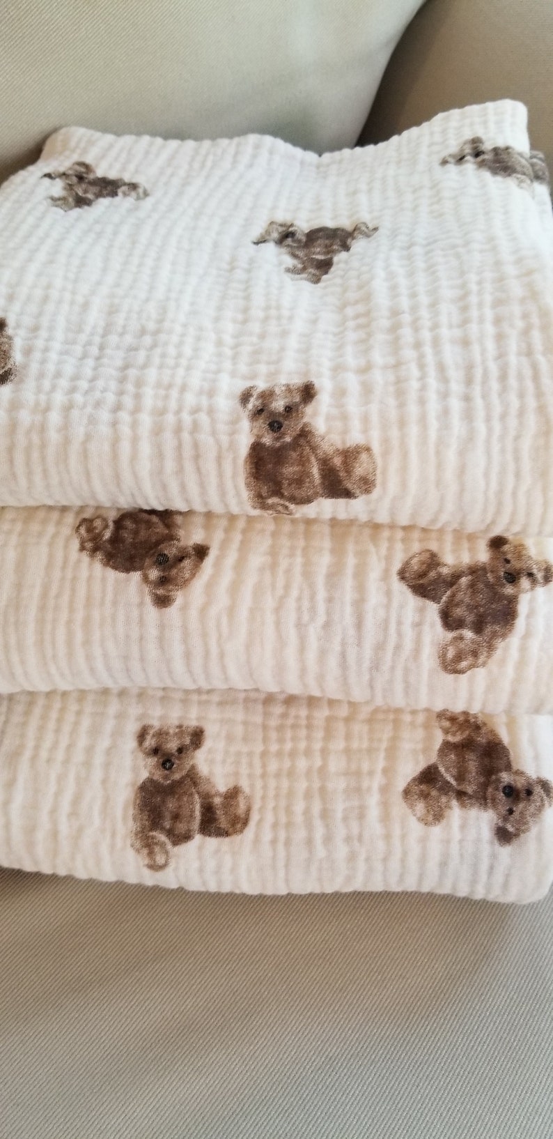 May include: A stack of three white muslin baby blankets with a brown teddy bear print. The blankets are soft and textured.