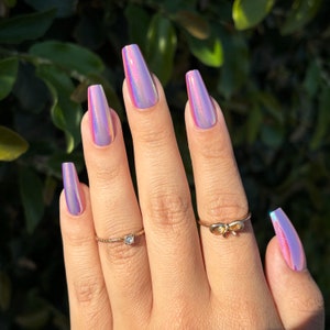 May include: A hand with long, coffin-shaped iridescent press-on nails. The nails reflect pink, purple, and gold colours. Two gold rings, one with a small diamond and the other with a bow design, are on the fingers.