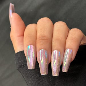 May include: Close-up of a hand with long, coffin-shaped artificial nails. The nails have a shimmering, iridescent finish with hues of pink, gold, and blue. The nails are a light, neutral color.