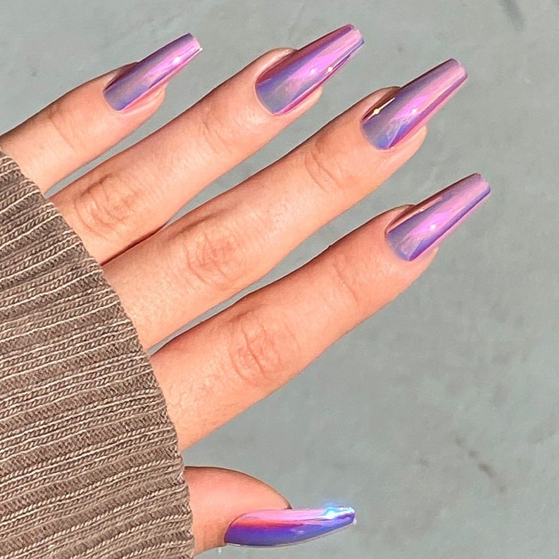 May include: A hand displaying long, coffin-shaped artificial nails. The nails have a holographic, iridescent finish, shifting between purple, pink, and blue. The nails are smooth and glossy, reflecting light.