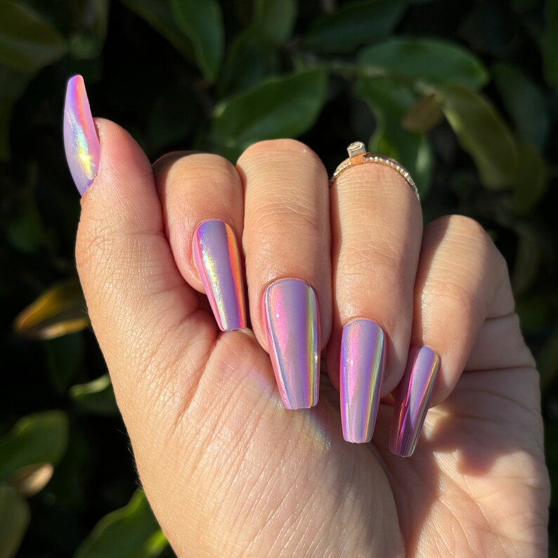 May include: A hand with long, coffin-shaped, iridescent nails. The nails shift in colour, displaying hues of pink, purple, and gold. A gold ring with a small diamond is on the ring finger.