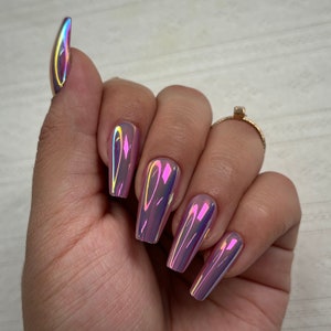 May include: A hand showing long, coffin-shaped artificial nails. The nails have a chrome finish, reflecting pink, purple, and yellow hues. A gold ring is visible on the ring finger. The nails are approximately 2 cm long.