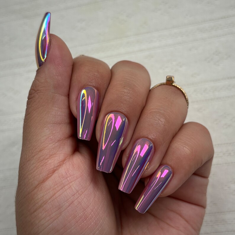 May include: A hand showing long, coffin-shaped artificial nails. The nails have a chrome finish, reflecting pink, purple, and yellow hues. A gold ring is visible on the ring finger. The nails are approximately 2 cm long.