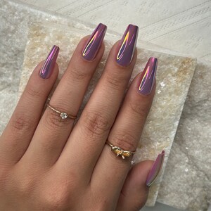 May include: A hand displaying long, coffin-shaped, iridescent purple nails. The nails reflect pink, yellow, and green hues. Two gold rings are worn: one with a small diamond and another with a bow design.