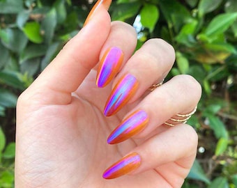 May include: Long, pointed fingernails with an iridescent, colour-shifting finish. The nails transition from orange to purple and blue, creating a vibrant effect. The hand also features gold rings.