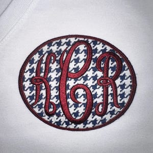May include: White t-shirt with an embroidered oval patch. The patch features a houndstooth pattern in navy blue and white, with a burgundy border. The letters "KR" are embroidered in burgundy script.