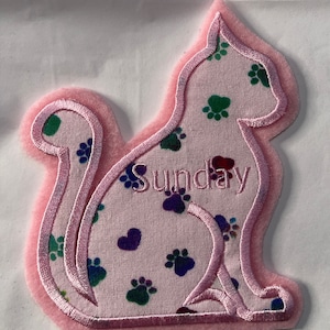 May include: Pink fabric cat-shaped patch with a pink border and a paw print pattern. The patch has the word "Sunday" embroidered on it.