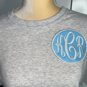 May include: A gray t-shirt with a blue circle containing a white embroidered monogram "H.G.R."