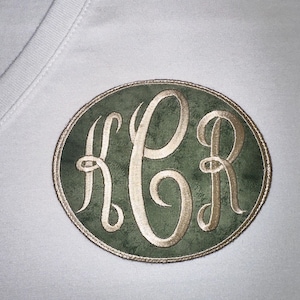 May include: An embroidered patch featuring the initials KCR in a flowing script. The letters are a light gold color, set against a textured green background within an oval shape. The patch is bordered with a gold trim and is affixed to a white fabric.
