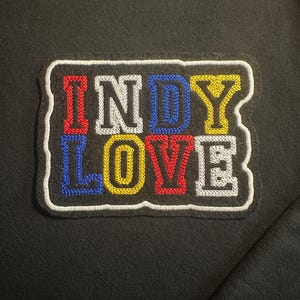 May include: Black sweatshirt with a colorful embroidered patch. The patch features the words "INDY LOVE" in red, blue, yellow, and white, with a white border. The letters are in a block style.