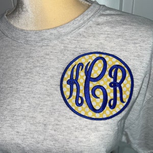 May include: A gray t-shirt with a blue and yellow monogram in a circle. The monogram is "HPR" with white polka dots on a yellow background.