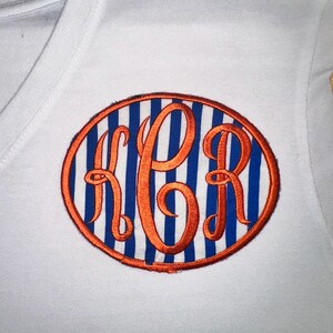 May include: White V-neck shirt with an embroidered oval patch. The patch features blue vertical stripes, an orange border, and orange monogram letters. The shirt is made of a soft, comfortable material.