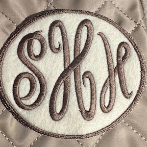 May include: An oval-shaped patch with a monogram in brown thread on a white background. The patch is surrounded by a brown border and set against a quilted, tan fabric.