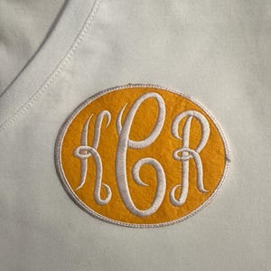 May include: A white V-neck shirt with an embroidered patch. The patch is an oval shape with a yellow background and white embroidered initials KCR. The patch is outlined with a white border.