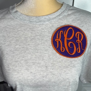 May include: A gray t-shirt with a purple and orange embroidered monogram patch. The monogram is in a circle and the letters are "KPB".