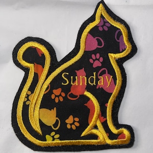 May include: A black fabric cat-shaped patch with a gold outline. The patch is decorated with a repeating pattern of colorful cats and paw prints. The word "Sunday" is embroidered in the center of the patch.