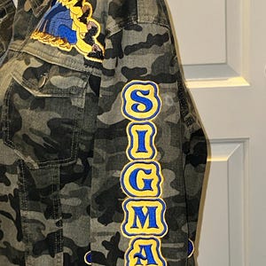 May include: Camouflage denim jacket with patches. The jacket features a large patch on the upper left chest and the word "SIGMA" in yellow and blue lettering down the left sleeve. Additional patches with text are visible.