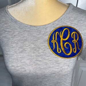 May include: A gray t-shirt with a blue and gold embroidered monogram in a circle.