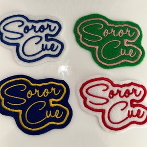 Puede incluir: Cuatro parches bordados con el texto "Soror Cue" en un esquema de colores blanco, verde, azul y rojo. Los parches tienen todos el mismo diseño, pero con diferentes colores.