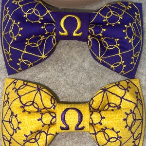 May include: Two bow ties with a gold and purple patterned design. The bow ties have a gold embroidered omega symbol in the center.