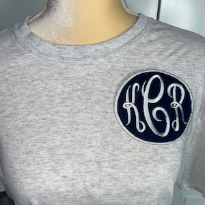 May include: A gray t-shirt with a navy blue and silver monogram patch. The monogram is in a circle and the letters are "KPB".
