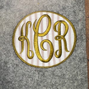 May include: An oval-shaped patch with a gold embroidered monogram of the letters KBR. The monogram is stitched in a cursive style and is centered on a white and tan striped background. The patch is outlined with gold thread.