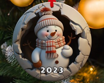 Puede incluir: Un adorno de muñeco de nieve blanco con una bufanda a rayas rojas y blancas y un sombrero negro. El adorno tiene forma de círculo y tiene un diseño de hielo agrietado alrededor de los bordes. El año 2023 está impreso en el adorno.