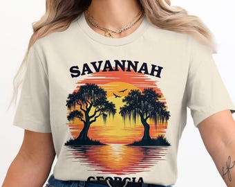 Puede incluir: Una camiseta beige con un estampado gráfico de una puesta de sol sobre un río con dos árboles. El texto "SAVANNAH GEORGIA" está impreso en la camiseta.