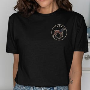 May include: A black short-sleeved t-shirt featuring a circular graphic on the left chest. The graphic depicts a dog silhouette filled with the American flag pattern, surrounded by three stars at the top and a heartbeat line at the bottom.