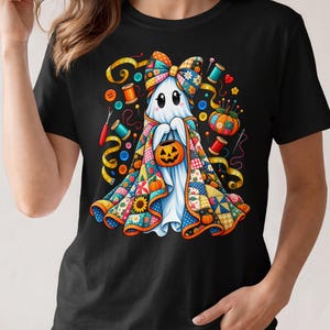 Könnte beinhalten: Ein schwarzes T-Shirt mit einer Grafik eines weißen Geistes, der eine große, bunte Patchwork-Decke und eine passende Schleife trägt. Der Geist hält einen kleinen orangefarbenen Kürbiseimer, umgeben von Nähzubehör wie Knöpfen, Garnrollen, Nadeln und einem gelben Maßband.