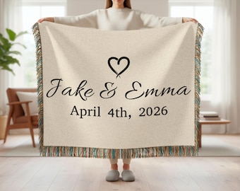 May include: A cream-colored throw blanket with a fringed edge, featuring the names "Jake & Emma" and the date "April 4th, 2026" in black script, with a heart symbol above the names. The blanket is held up by a person.