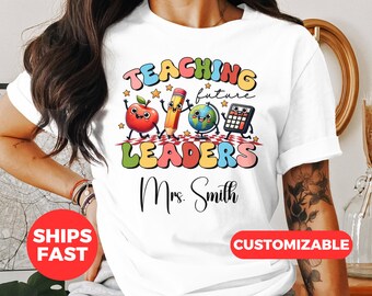 May include: White t-shirt with a colourful graphic that says "Teaching Future Leaders" with a cartoon apple, pencil, globe, and calculator. The text "Mrs. Smith" is below the graphic.