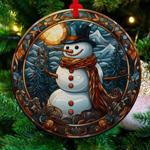 Peut inclure: Une boule de Noël ronde représentant une illustration de style vitrail d'un bonhomme de neige portant un chapeau haut de forme bleu et une écharpe brune. Le bonhomme de neige se tient dans un paysage enneigé avec des arbres et une pleine lune en arrière-plan.
