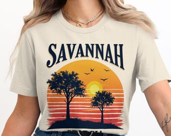 Puede incluir: Camiseta color crema con un diseño de puesta de sol y la palabra "SAVANNAH" en escritura azul marino. El gráfico incluye siluetas de árboles, pájaros y una vibrante puesta de sol naranja y amarilla.