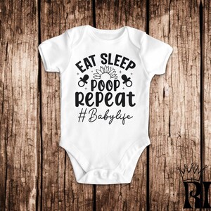 以下が含まれることがあります： 白いベビーロンパースに、黒い文字で「Eat Sleep Poop Repeat #Babylife」と書かれています。テキストの周りは、小さな太陽と哺乳瓶のグラフィックで囲まれています。