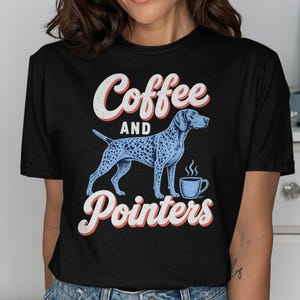 Può includere: Una t-shirt nera a maniche corte con una stampa grafica di un cane pointer blu a pois in piedi accanto a una tazza di caffè blu fumante. Il testo "Coffee AND Pointers" è stampato in un carattere bianco in stile retrò con bordo rosso.