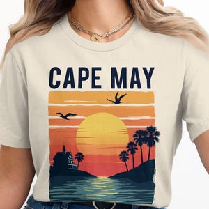 Puede incluir: Una camiseta beige con un estampado gráfico de una puesta de sol sobre una playa con palmeras y una casa. El texto "CAPE MAY" está impreso en negro sobre el gráfico.