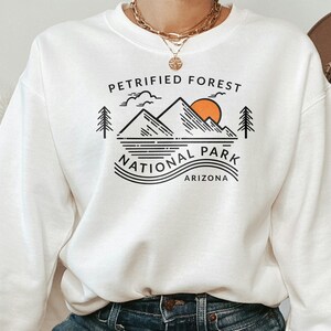 May include: White sweatshirt with a graphic design featuring the text "PETRIFIED FOREST NATIONAL PARK ARIZONA". The design includes mountains, a sun, trees, and water lines, all in black and orange.