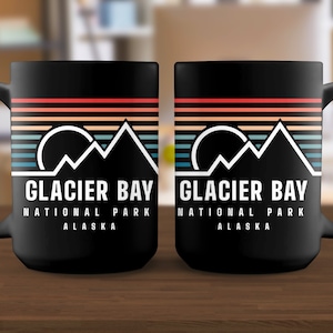 May include: Two black ceramic mugs with a retro-style design featuring a mountain range and a sun. The text on the mugs reads "Glacier Bay National Park Alaska".