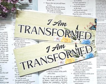 May include: Two rectangular bookmarks with the text "I Am TRANSFORMED" in a stylized font, adorned with floral and butterfly illustrations. The bookmarks rest on an open book with biblical text.